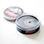 An open tin of Clay | Uppercut Deluxe, a strong hold hair styling clay by Uppercut Deluxe, with the lid behind the container on a white surface.