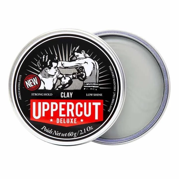 An open tin of Uppercut Deluxe Clay reveals its gray, matte-finish hair product and the labeled black, red, and white lid featuring a boxing illustration.