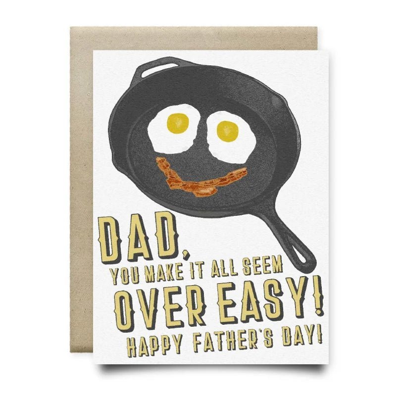 Anvil Cards Dad Makes It Over Easy by Anvil Cards is an A2 greeting card showcasing a frying pan with two sunny-side-up eggs and bacon arranged as a smiling face, accompanied by the message: “Dad, you make it all seem over easy! Happy Father’s Day!” Printed on premium linen paper and blank inside for your personal message.