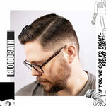 Man with brown hair, beard, and glasses sports a classic side-parted haircut styled with Deluxe Pomade by Uppercut Deluxe. Bold "BLOODBATH" and other cut-out words frame the image.
