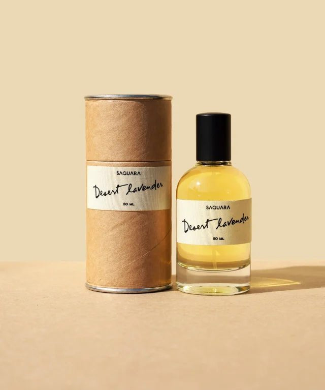 A bottle of Desert Lavender by Saguara Perfumes rests beside its cylindrical cardboard box against a beige background.