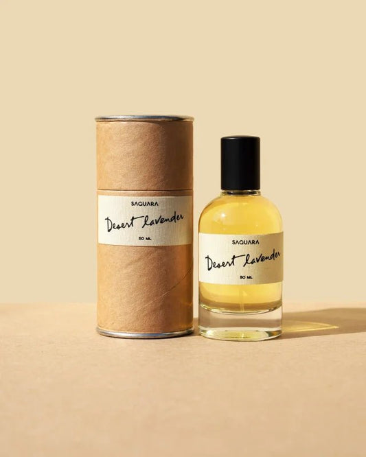 A bottle of Desert Lavender by Saguara Perfumes rests beside its cylindrical packaging on a beige surface, showcasing the California-made scent’s desert lavender allure.
