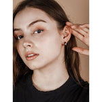 A young woman with long brown hair touches her ear, revealing JaxKelly’s Wide Hoop Earrings. She wears a black shirt and looks directly at the camera against a beige background.