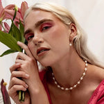 Woman with platinum blonde hair holds pink lilies, wearing bold pink eyeshadow, the Edith Necklace by Larissa Loden—a freshwater pearl necklace with gold fill beads—and pearl earrings, against a neutral background.