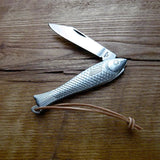 The Mollyjogger Fingerling Fish Knife features a silver, fish-shaped design with a textured body, short stainless blade, leather loop, and is shown lying on a wooden surface.