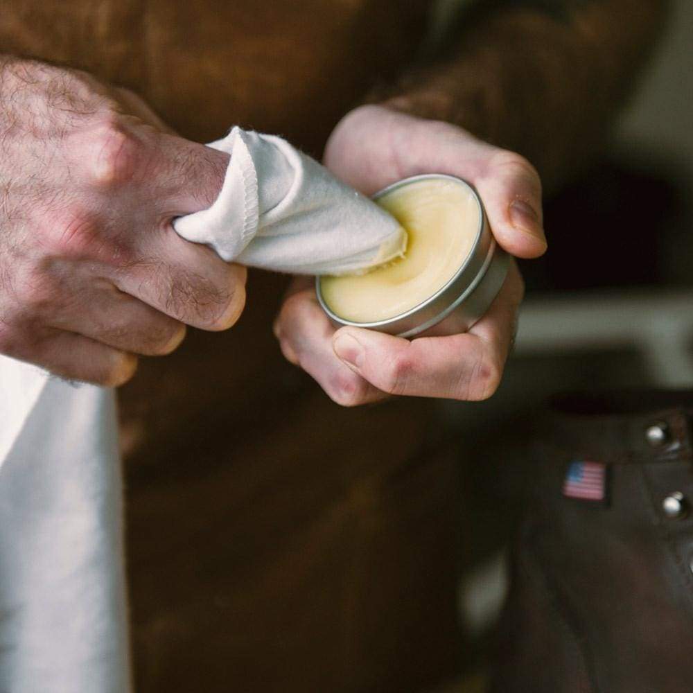 Someone uses the Otter Wax Flannel Buffing Cloth to pick up balm or polish, ready to apply it to a leather boot with an American flag patch.