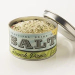 An open tin of Beautiful Briny Sea French Picnic Sea Salt, filled with a coarse blend of Pure Ocean Salt and herbes de Provence, sits on a white surface with the lid beside it.
