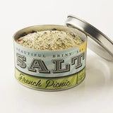 An open tin of Beautiful Briny Sea French Picnic Sea Salt, filled with a coarse blend of Pure Ocean Salt and herbes de Provence, sits on a white surface with the lid beside it.