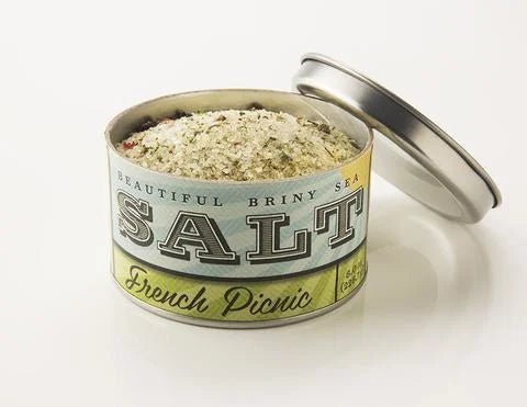 An open tin of Beautiful Briny Sea French Picnic Sea Salt, filled with a coarse blend of Pure Ocean Salt and herbes de Provence, sits on a white surface with the lid beside it.