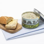 A wooden board with sliced bread sits beside an open tin of French Picnic Sea Salt by Beautiful Briny Sea, placed on a folded blue and white cloth.
