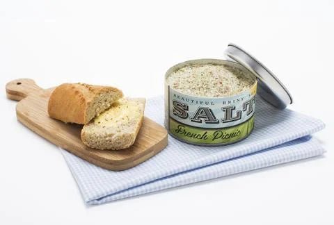 A wooden board with sliced bread sits beside an open tin of French Picnic Sea Salt by Beautiful Briny Sea, placed on a folded blue and white cloth.