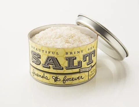 An open tin of "Friends Forever Sea Salt" by Beautiful Briny Sea, filled with coarse salt. The label suggests a salty-sweet blend, combining sea salt with honey and the phrase "friends forever.