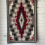 The Vintage Ganado Rug Classic "Snowflake" CA 1980, featuring geometric red, black, white, and gray patterns, hangs on a white wooden wall and showcases traditional Navajo weaving.