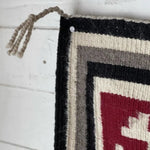 Close-up of a Vintage Ganado Rug Classic "Snowflake" CA 1980, featuring black, gray, cream, and red geometric patterns, pinned to a white wooden wall by a nail in the upper left corner.