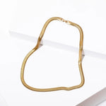 The Grace Necklace by Larissa Loden, a flat gold snake chain, is displayed on a white, angled surface.