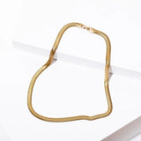 The Grace Necklace by Larissa Loden, a flat gold snake chain, is displayed on a white, angled surface.
