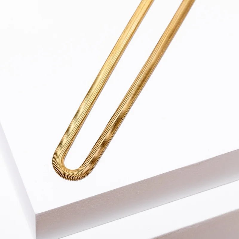 A close-up of the Grace Necklace by Larissa Loden, featuring a gold-colored, textured metal rod bent into a U-shape, resting on the edge of a white rectangular surface.