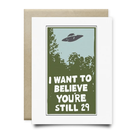The "I Want to Believe You're Still 29 Card" by Anvil Cards features a UFO illustration above trees with the caption "I WANT TO BELIEVE YOU'RE STILL 29," printed on linen paper, blank inside, with brown envelope included.