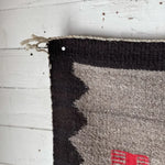 The Vintage Klagetoh Rug Circa 1950 with black, gray, and red patterns hangs on a white wooden wall, secured by a nail at the top corner.