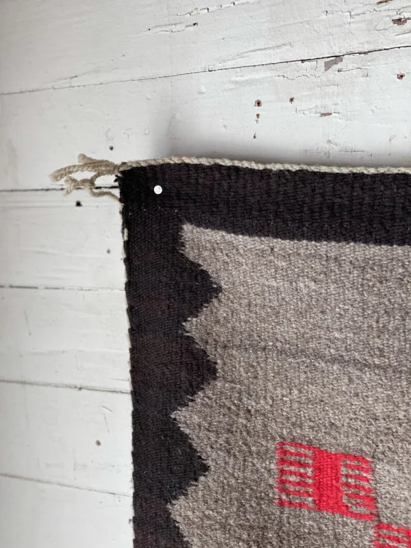 A close-up of a Klagetoh Rug Circa 1950 by Vintage, showing black, gray, and red patterns in the corner, pinned to a white wooden floor.