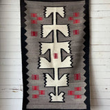 A Vintage Klagetoh Rug Circa 1950 with geometric black, white, gray, and red patterns hangs on a white wooden wall.