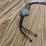 The Vintage Large Turquoise Bolo features a prominent turquoise stone and braided black leather cord, displayed on a wooden surface.