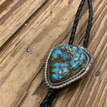The Vintage Large Turquoise Bolo features a braided black leather cord and a prominent Kingman turquoise stone in a decorative silver frame, displayed on a wooden surface.