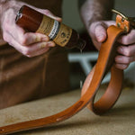 A person uses both hands to apply Otter Wax Leather Polishing Oil from a bottle onto a brown leather belt over a work surface.