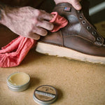 A person polishes a brown leather boot with a red cloth as an open tin of Otter Wax Leather Salve sits nearby, ready to provide deep conditioning.