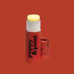 A tube of Poppy & Pout Lip Balm with the cap off reveals the balm inside. The red packaging bears black text and floral designs, displayed against a matching red backdrop.