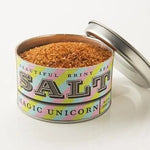 An open tin of Beautiful Briny Sea Magic Unicorn Sea Salt, with a pastel label and detached lid nearby, holds coarse, brownish ocean salt.