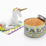 An open tin of Magic Unicorn Sea Salt by Beautiful Briny Sea sits beside a white ceramic unicorn on a colorful chevron napkin, capturing whimsy and suggesting hints of rosemary and ocean salt inside.