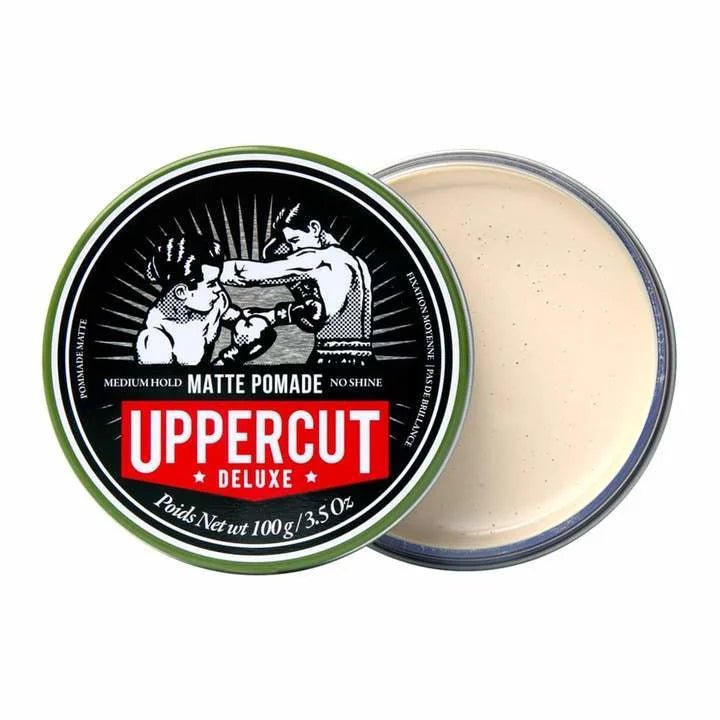 A round container of Uppercut Deluxe Matte Pomade with a partially removed lid reveals the cream-colored product inside. The lid displays a vintage boxing illustration, highlighting medium hold and a natural finish.