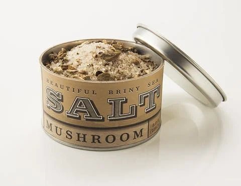 An open tin of Beautiful Briny Sea’s Mushroom Sea Salt, filled with a coarse, umami-rich salt blend made with foraged mushrooms, rests on a white surface with its lid leaning to the side.