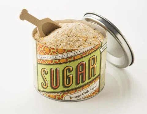 An open tin of Beautiful Briny Sea’s Orange Chili Sugar, with organic cane sugar and orange clementine peel, displays a wooden scoop nestled inside as the lid sits beside it.