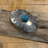 The Oval Sterling Silver w/ Turquoise Brooch by Vintage features decorative carving and a central turquoise cabochon, elegantly displayed on weathered wood.
