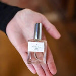 A hand holds a small glass bottle of Parfum | Rita’s Car by Saint Rita Parlor, featuring a silver cap and white label, showcasing this handmade fragrance crafted from organic essential oils.