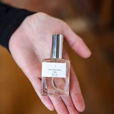 A hand holds a small glass bottle of Parfum | Rita’s Car by Saint Rita Parlor, featuring a silver cap and white label, showcasing this handmade fragrance crafted from organic essential oils.