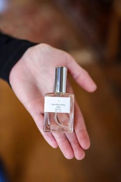 A hand holds a small glass bottle of Parfum | Rita’s Car by Saint Rita Parlor, featuring a silver cap and white label, showcasing this handmade fragrance crafted from organic essential oils.