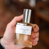 A hand holds a clear glass perfume bottle with a silver cap. The white label reads “Parfum | Rita’s Car | Saint Rita Parlor,” showcasing this unisex fragrance by Saint Rita Parlor, crafted with organic essential oils.