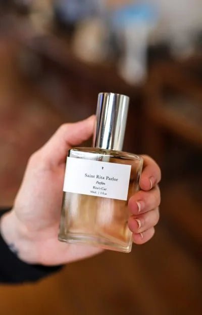 A hand holds a clear glass perfume bottle with a silver cap. The white label reads “Parfum | Rita’s Car | Saint Rita Parlor,” showcasing this unisex fragrance by Saint Rita Parlor, crafted with organic essential oils.