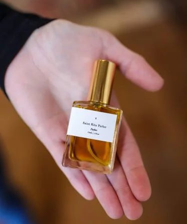 A hand holds a small square glass bottle of Parfum | Signature | Saint Rita Parlor by Saint Rita Parlor, featuring a gold cap and white label, highlighting this unique unisex fragrance.