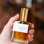 A hand holds a rectangular glass perfume bottle with a gold cap and a white label reading "Saint Rita Parlor," featuring the Signature Parfum, a handmade unisex fragrance by Saint Rita Parlor.