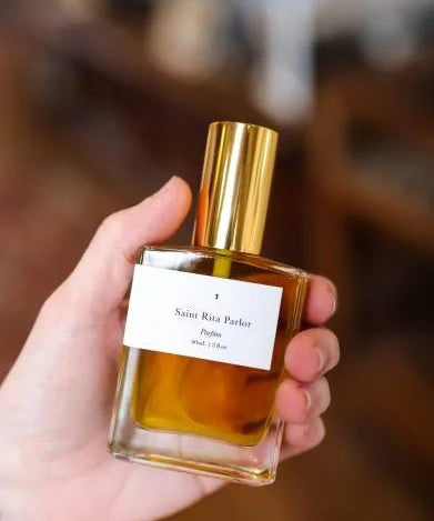 A hand holds a glass perfume bottle with a gold cap and a white label reading "Saint Rita Parlor." The featured unisex scent is Parfum | Signature by Saint Rita Parlor, shown in focus against a blurred background.