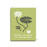 The "Peace on Your Birthday Card" by Worthwhile Paper is a green recycled card featuring white lotus blossoms, lily pads, and the message "Wishing you peace on your birthday" printed on the front.