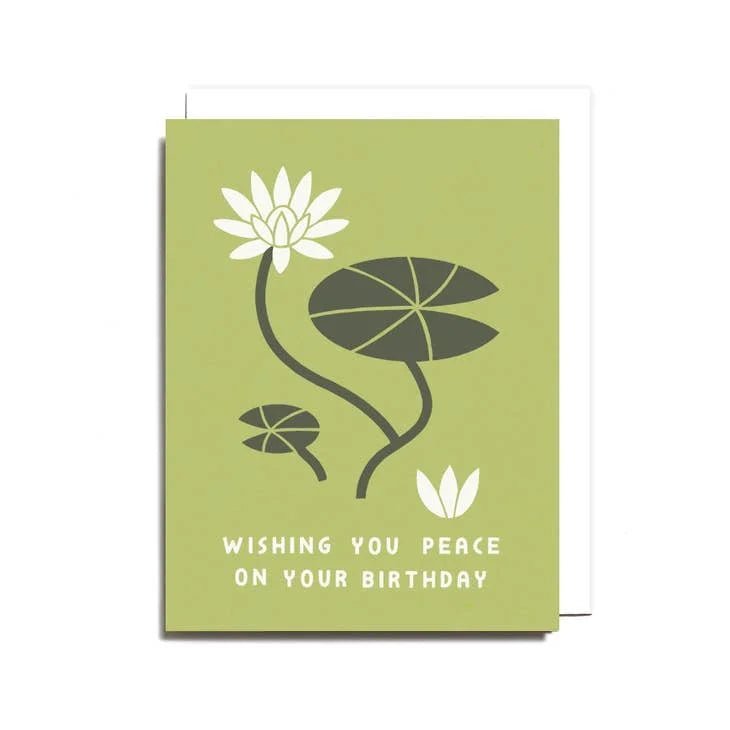 The "Peace on Your Birthday Card" by Worthwhile Paper is a green recycled card featuring white lotus blossoms, lily pads, and the message "Wishing you peace on your birthday" printed on the front.