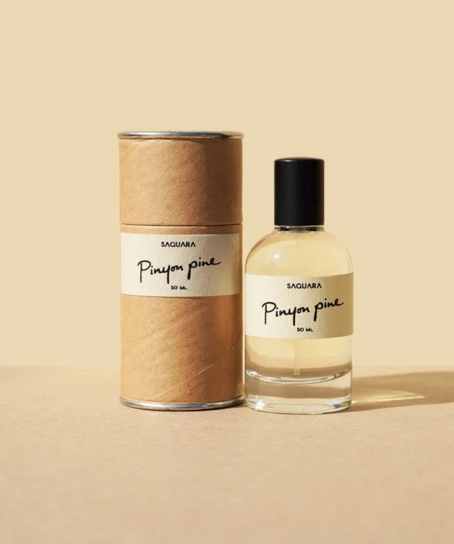 A bottle of Pinyon Pine by Saguara Perfumes, handmade in California, is displayed beside its cylindrical cardboard packaging against a beige background.
