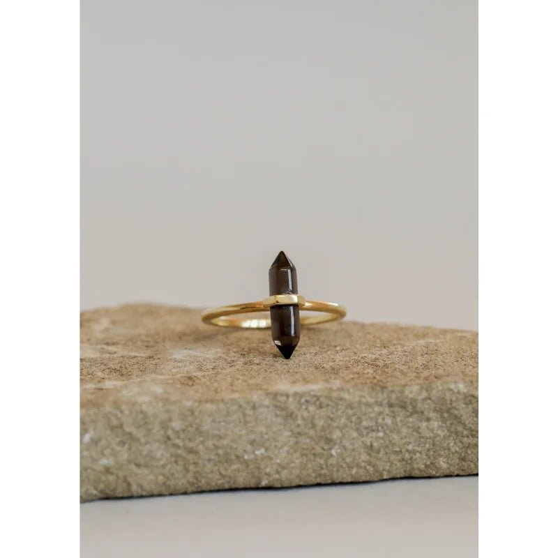 The JaxKelly Ring | Smokey Quartz Point features a hand-crafted gold vermeil band with a double-pointed dark brown crystal, displayed upright on a stone surface against a plain background.