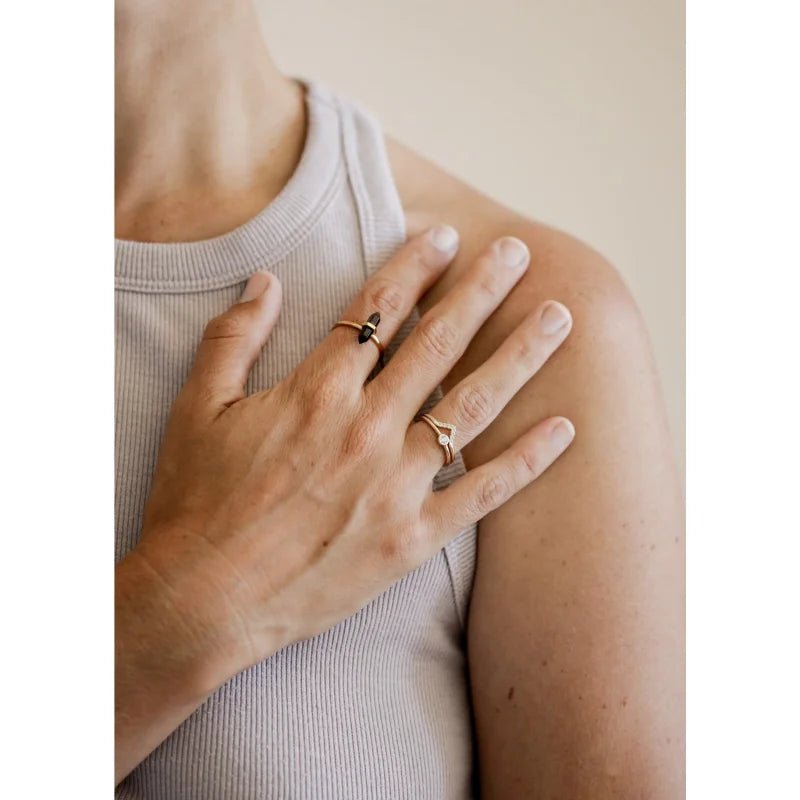 A person in a ribbed sleeveless top rests their hand on their shoulder, showcasing two handcrafted gold rings from JaxKelly, including the Ring | Smokey Quartz Point | JaxKelly adorned with gemstones.