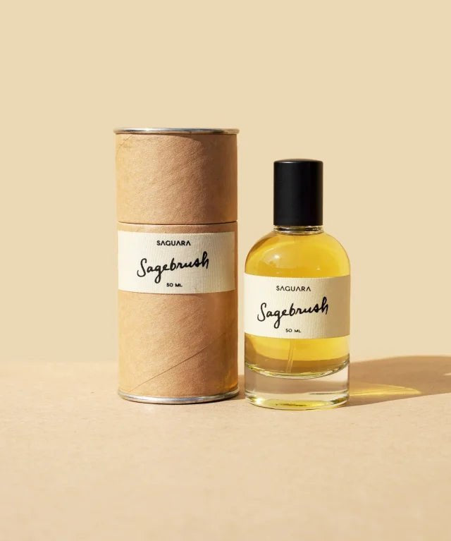 A clear glass bottle labeled "Sagebrush | Saguara Perfumes" (50 mL Eau de Parfum, vegan, with California sage notes) and its cylindrical cardboard container from Saguara Perfumes are displayed on a beige surface and background.